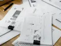 Stos papierów z wykresami i danymi, spinacze biurowe, długopis na drewnianym biurku.