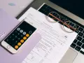 Telefon z kalkulatorem, okulary i dokumenty podatkowe na laptopie.